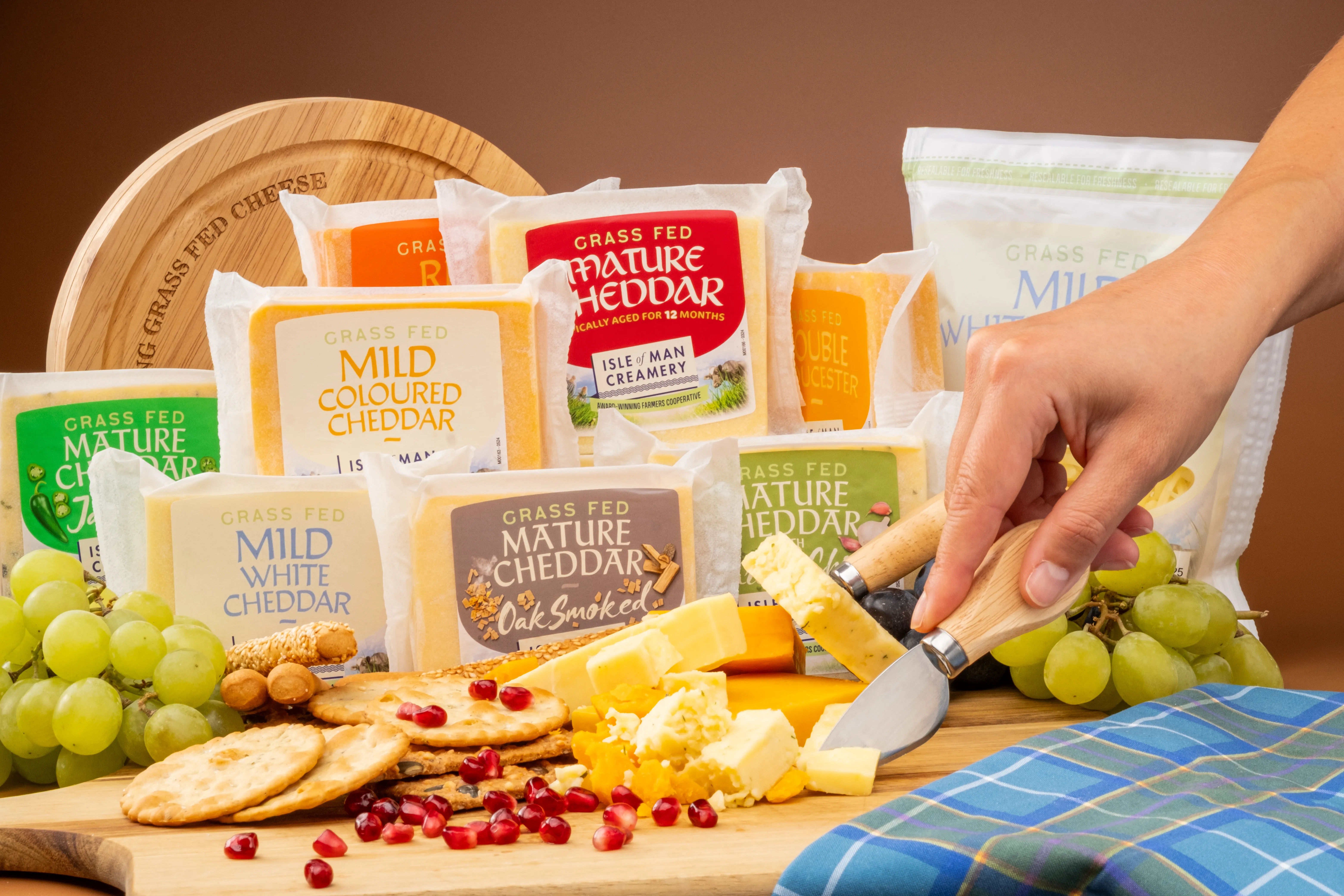 A hand slicing into a wedge of Isle of Man Creamery mature cheddar on a wooden board, surrounded by a range of grass-fed cheddar products including mild, mature, coloured, white and oak smoked varieties, with crackers, grapes and pomegranate seeds